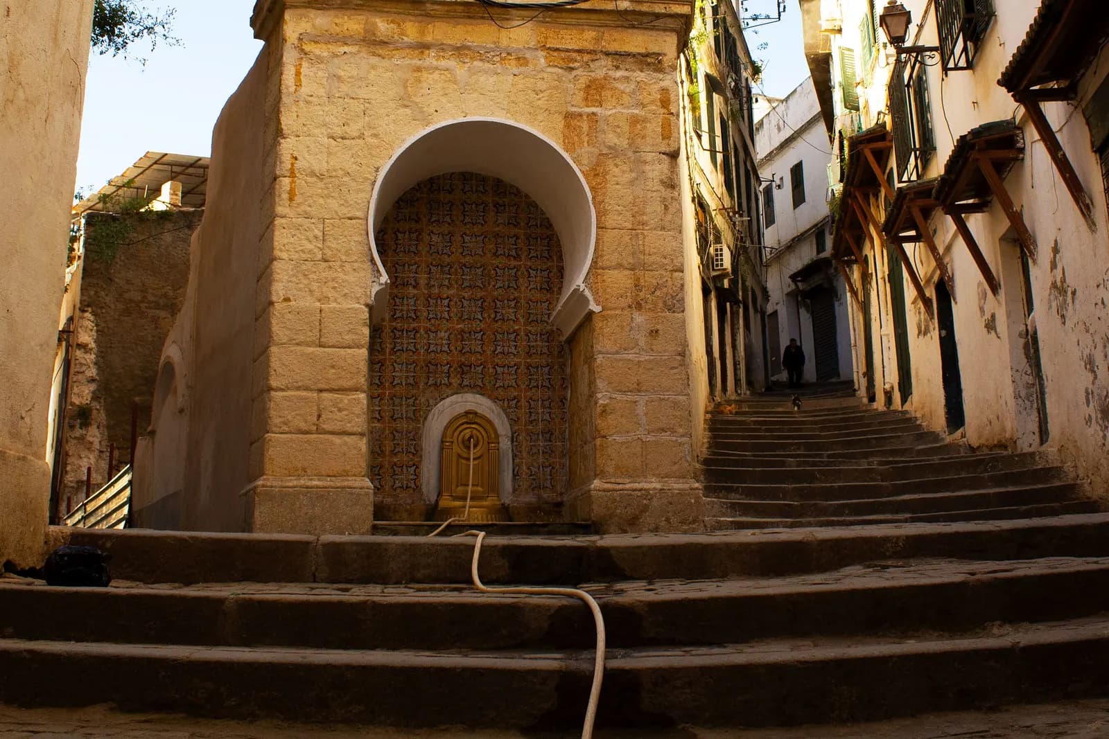 Une ruelle en escalier de la Casbah d’Alger et sa fontaine sous un arc outrepassé.